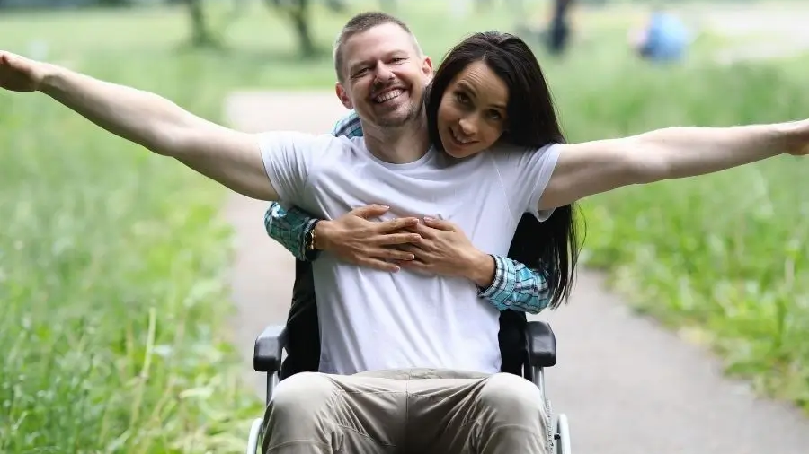 Rollstuhlfahrer wird von hinten von seiner Freundin umarmt. Er hat einen Rollstuhl gemietet und sie machen einen Ausflug auf einer Wiese. 